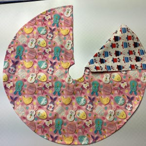 May include: A pink and white fabric tree skirt with a pastel Easter egg and bunny pattern. The skirt has a white fabric center with a red, white, and blue cake pattern.