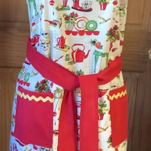 May include: A red and white apron with a kitchen-themed print. The apron has a red tie and two red pockets with yellow scalloped edges.