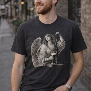 Man standing on a city street wearing a faded black vintage-style graphic T-shirt featuring an angel pouring a pint, looking to the side with a relaxed expression.