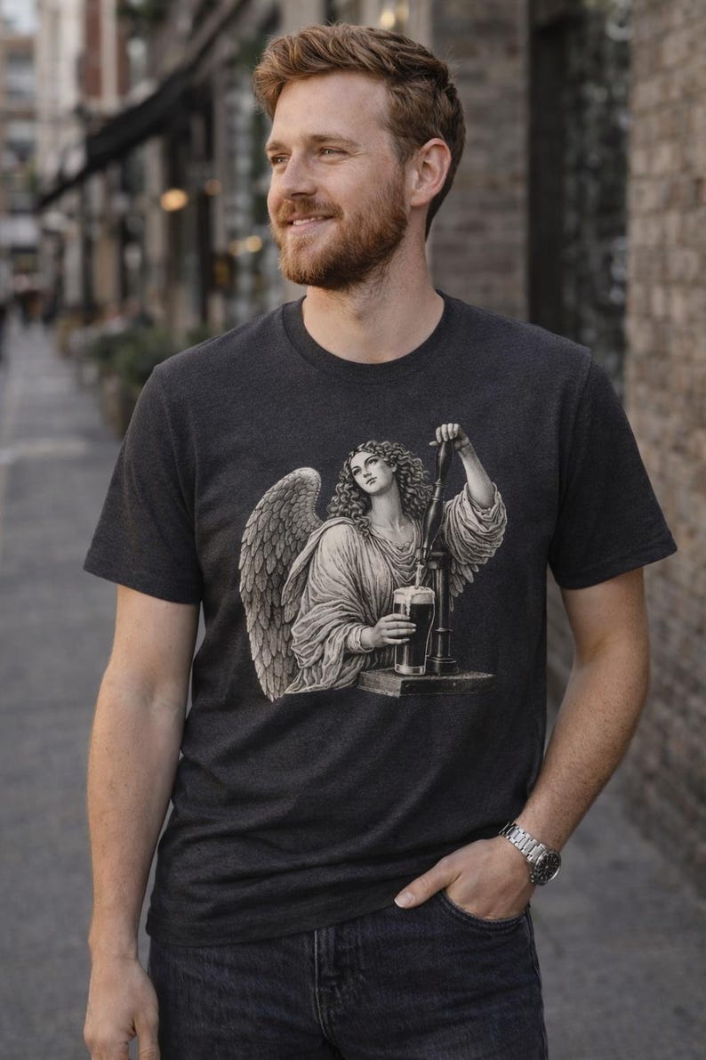 Man standing on a city street wearing a faded black vintage-style graphic T-shirt featuring an angel pouring a pint, looking to the side with a relaxed expression.