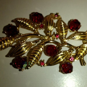 Könnte beinhalten: Goldfarbene Brosche mit botanischem Design. Sie zeigt Blätter und Beeren, besetzt mit roten und rosa Edelsteinen. Das detaillierte, strukturierte Design erinnert an ein Blumenarrangement, ideal, um jedem Outfit einen Hauch von Eleganz zu verleihen.