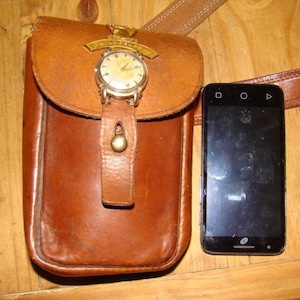 Puede incluir: Una bolsa de cuero marrón con un reloj dorado adjunto. El reloj tiene una esfera blanca y agujas negras. La bolsa cuelga de una correa de cuero marrón. Un teléfono inteligente negro está sentado sobre una superficie de madera junto a la bolsa.