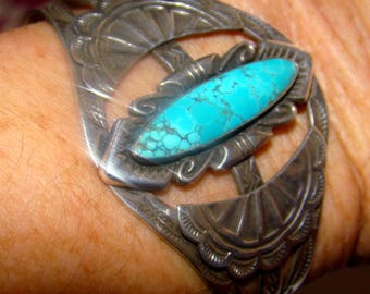 May include: A silver cuff bracelet featuring a central marquise-shaped turquoise stone with dark matrix veining. The metal band displays intricate stamped fan-like patterns on both sides of the stone, creating a wide, open-work design.