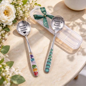 May include: Two silver-toned metal soup spoons with engraved text reading Jason's Soup Spoon. One spoon features a heart icon, the other a smiley face. The handles are decorated with colorful, square, glittery inlays. A clear plastic case with a green ribbon is included.