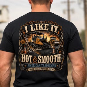 May include: A black t-shirt featuring a graphic on the back of an asphalt paver and road roller at sunset. The text reads I LIKE IT HOT & SMOOTH, AMERICAN TRADESMAN, and BLUE COLLAR ASPHALT CREW, framed by a chain border with orange traffic cones.