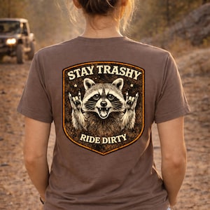 Può includere: Maglietta marrone con una grafica di un procione che fa il gesto del rock and roll. Il testo sulla maglietta dice "STAY TRASHY" sopra il procione e "RIDE DIRTY" sotto. La maglietta viene indossata all'aperto.