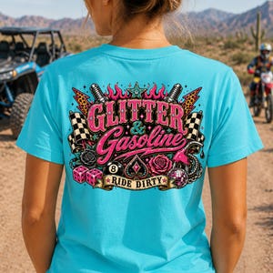 Op de afbeelding: Turquoise t-shirt met een roze en zwart grafisch ontwerp op de achterkant. Het ontwerp bevat de woorden "Glitter & Gasoline", vlammen, een ster, dobbelstenen, rozen en een geblokte vlag. De woorden "Ride Dirty" zijn ook opgenomen.