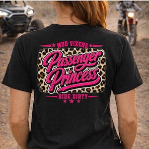 May include: A black t-shirt featuring a graphic on the back with a leopard print rectangular background. The text reads "MUD VIXENS" in pink, "Passenger Princess" in large pink script, and "RIDE DIRTY" in pink, accented with pink stars.
