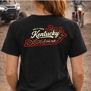 May include: A black t-shirt featuring a graphic on the back of the outline of the state of Kentucky filled with a brown and black leopard print pattern. The text reads "SHE'S FROM Kentucky I can tell" in white and cream script and block lettering.