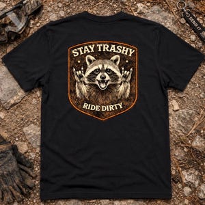 May include: A black short-sleeved t-shirt featuring a central graphic of a raccoon making a rock-and-roll hand gesture. The design is enclosed in a shield-shaped border with the text STAY TRASHY at the top and RIDE DIRTY at the bottom in white.