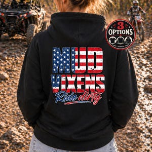 May include: Black hoodie with the text "MUD VIXENS Ride dirty" in a patriotic design. The text features the American flag colors and stars. The hoodie has a drawstring hood and a front pocket. The image also shows a UTV and a dirt bike.
