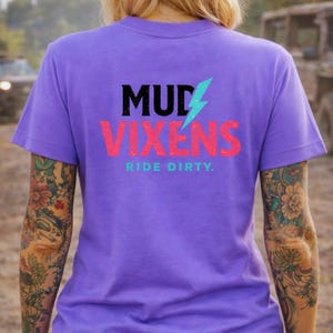May include: A purple t-shirt featuring the text "MUD VIXENS" in black and pink, with a blue lightning bolt graphic. The words "RIDE DIRTY" are printed below. The shirt has short sleeves.
