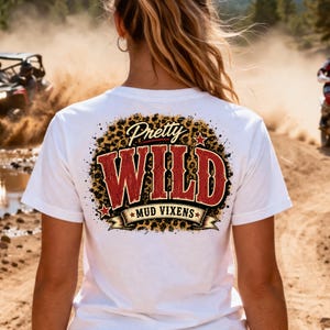 May include: A white t-shirt featuring a graphic on the back with a leopard print background. The text reads Pretty WILD in large red letters, with MUD VIXENS printed on a banner below. The design includes small red stars and a distressed texture.