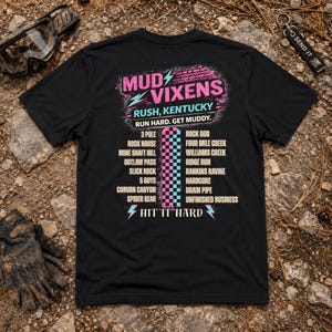 Op de afbeelding: Een zwart t-shirt met korte mouwen en een grafische print op de achterkant. Het ontwerp bevat de tekst MUD VIXENS, RUSH, KENTUCKY, RUN HARD. GET MUDDY. en HIT IT HARD, naast een lijst met locaties en een centraal roze en blauw dambord.