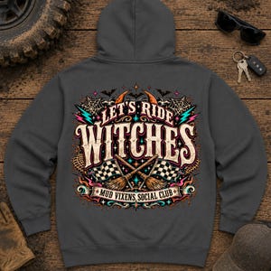 Op de afbeelding: Een antracietgrijze hoodie met een graphic op de achterkant met de tekst "LET'S RIDE WITCHES" en "MUD VIXENS SOCIAL CLUB". Het ontwerp bevat gekruiste bezems, geblokte vlaggen, spinnenwebben, vleermuizen en kleurrijke sterren.
