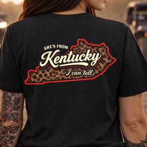 Może przedstawiać: Czarny t-shirt z napisem "She's From Kentucky I can tell". Słowo "Kentucky" jest w stylizowanej czcionce z wypełnieniem w panterkę, obrysowane na czerwono. Odzież codzienna.