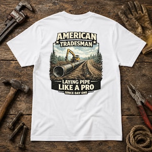 May include: A white t-shirt featuring a graphic on the back with the text AMERICAN TRADESMAN, LAYING PIPE LIKE A PRO, SINCE DAY ONE. The illustration depicts a yellow excavator working on a large metal pipe in a forest setting with dirt.