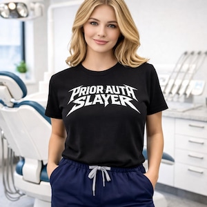 以下が含まれることがあります： 白い文字「PRIOR AUTH SLAYER」が入った黒いクルーネックTシャツ。ネイビーブルーのスクラブと合わせています。画像は歯科医院で撮影されました。