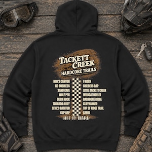 Op de afbeelding: Een zwarte hoodie met een afbeelding op de achterkant met de tekst TACKETT CREEK HARDCORE TRAILS, een lijst met trailnamen, een centrale geblokte kolom en de tekst HIT IT HARD, tegen een textuurrijke achtergrond met modderspatten.