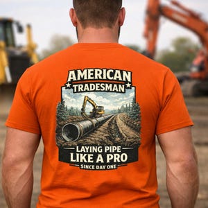 May include: Orange t-shirt featuring the text "AMERICAN TRADESMAN" above a construction scene illustration with a backhoe laying pipe. The text "LAYING PIPE LIKE A PRO SINCE DAY ONE" is below the illustration.
