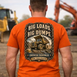 May include: An orange t-shirt featuring a graphic on the back. The design shows a yellow dump truck, a hard hat, gloves, and a wrench inside a chain-link border. Text reads: BIG LOADS. BIG DUMPS. AMERICAN TRADESMAN DUMP TRUCK OPERATOR.