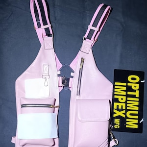 May include: A pink leather chest bag with black and white striped straps. The bag has multiple zippered pockets and a small pouch. A black and yellow tag with the text "OPTIMUM IMPEX MFG" is visible.