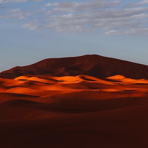 May include: A scenic landscape of a desert at sunset. The image features rolling sand dunes in shades of orange and red, with a mountain in the background. The sky is a gradient of blue and white, with a few clouds.