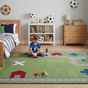 May include: A child's bedroom with a green road-themed rug. The rug features a grey road design with red houses, blue crosses, and trees. A wooden bed, chest of drawers, and storage chest are also in the room. Footballs and a football wall decoration are also visible.
