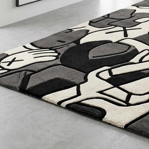 May include: A rectangular area rug with a modern design. The rug features a pattern of abstract shapes in shades of gray, black, and white. The design includes stylized hands and geometric forms, creating a contemporary aesthetic. The rug is placed on a light gray floor.