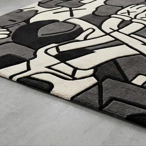May include: A rectangular rug with a modern, abstract design in shades of black, gray, and white. The rug features a geometric pattern with thick black outlines and a plush texture. The design includes stylized hands and shapes.