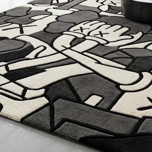 May include: A rectangular rug with a geometric design in shades of gray, black, and white. The rug features a stylized cartoon figure with crossed arms and hands with an "X" on each. The rug is made of a soft, plush material.