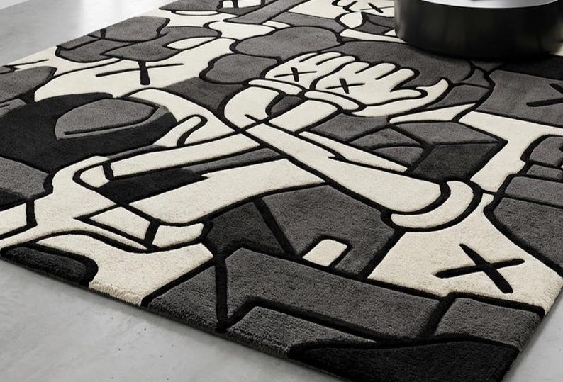 May include: A rectangular rug with a geometric design in shades of gray, black, and white. The rug features a stylized cartoon figure with crossed arms and hands with an "X" on each. The rug is made of a soft, plush material.