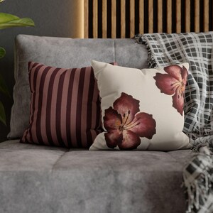 Op de afbeelding: Twee decoratieve sierkussens op een grijze bank. Eén kussen heeft verticale bordeauxrode strepen. Het andere kussen is crèmekleurig met twee grote bordeauxrode bloemmotieven. Een geruite deken is op de achtergrond gedrapeerd.