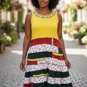 May include: A sleeveless, patchwork dress with a yellow top and a skirt featuring red, green, and floral-print panels. The dress has a round neckline with a decorative trim and a ruffled hem. The woman is standing on a cobblestone street.