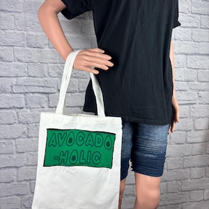 May include: A white tote bag with the words "AVOCADO -HOLIC" in green block letters on a green rectangle. The bag is held by a person wearing a black t-shirt and blue denim shorts.