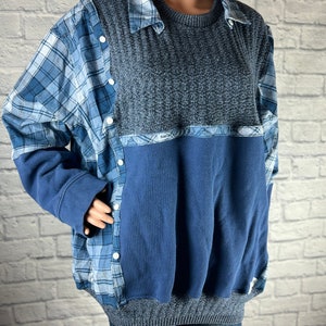 May include: A blue and grey sweater with a plaid shirt layered on top. The sweater has a ribbed texture and a blue and white plaid pattern on the bottom.