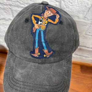 May include: A charcoal gray baseball cap featuring an embroidered patch of the character Woody from the Toy Story movie. The patch shows Woody in his signature cowboy outfit. The cap has a curved brim and a fabric-covered button on top.