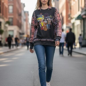 May include: A long-sleeve sweatshirt featuring a dark grey centre panel with a colourful Scooby-Doo graphic print, accented by pink and black plaid fabric on the sleeves and hem, paired with blue denim jeans and white trainers.