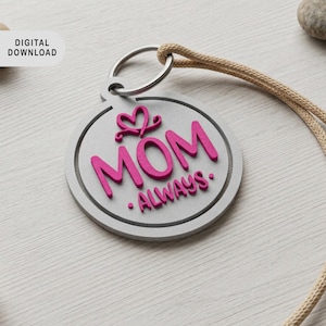 Pode incluir: Um chaveiro redondo cinza com a palavra "MOM" em rosa brilhante, junto com "ALWAYS". Um design de coração e coroa rosa está acima da palavra "MOM". O chaveiro tem um anel prateado e um cordão bege.