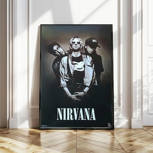May include: A rectangular framed poster featuring a black and white photograph of the band Nirvana. The image shows three members in a studio setting, with the word NIRVANA printed in large, bold, white capital letters at the bottom of the frame.