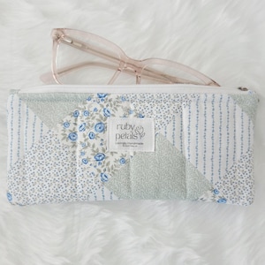 May include: A glasses case with a floral pattern and a pair of pink-framed glasses. The case has a white zip and a label that reads "ruby petals Loving Handmade". The case is white with blue floral accents.