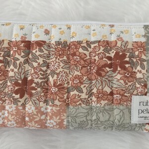 May include: A rectangular quilted zip pouch featuring a patchwork design of floral patterns in shades of terracotta, sage green, and cream. A white fabric label on the bottom right corner displays the text ruby petals Lovingly Handmade.