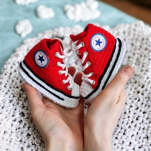 May include: A pair of red and white crochet baby sneakers with white laces and a blue star emblem. The sneakers have a black stripe along the white sole. The shoes are held in open hands.