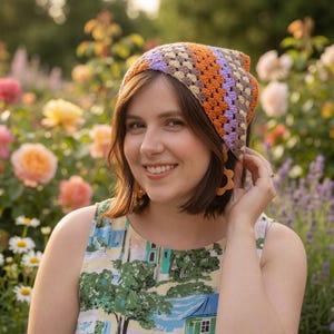 Peut inclure: Un couvre-chef au crochet coloré dans les tons orange, violet et beige. Le couvre-chef est porté par une personne dans une robe à motifs floraux, souriant dans un jardin. Le couvre-chef est de forme carrée, avec un motif granny square.