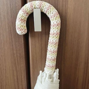 Può includere: Un copri manico per ombrello all'uncinetto con un motivo in filato variegato in delicate tonalità di rosa, giallo e bianco. La copertura ha la forma di un bastoncino e avvolge il manico curvo, con il tessuto bianco dell'ombrello visibile sotto.