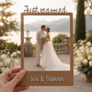 Könnte beinhalten: Ein rechteckiger, hellbrauner Holz-Fotorahmen mit dem weißen Schriftzug Just married. oben und dem weißen Schriftzug lea & Fabian unten. Der Rahmen ist so gestaltet, dass er gehalten werden kann, um ein Paar in der Ferne einzurahmen.