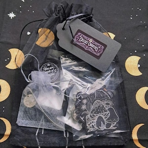 May include: A black drawstring bag with a tag reading "Occult Oddities." The bag contains charms, a mushroom pendant, and other items in small bags and clear packaging. The background has a celestial design with moons and stars.