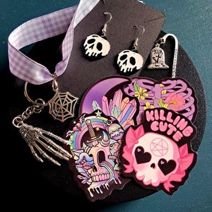 Puede incluir: Una colección de accesorios de temática gótica, que incluye pendientes de calavera, un llavero de telaraña y pegatinas con diseños de calaveras y arcoíris. También hay una cinta de cuadros morada y un marcapáginas con una lápida.