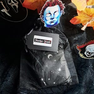Op de afbeelding: Een zwarte cadeauzak met een label met de tekst "Slasher Stash". De zak is versierd met een hemels patroon van sterren en manen. Decoratieve uitsparingen van horrorfilmkarakters zijn ook aanwezig.