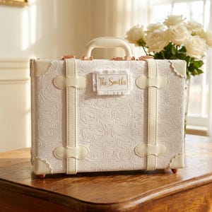 May include: A white suitcase with a floral pattern, cream-colored straps, and a handle. The suitcase has the text "The Smiths" on a small label. It sits on a wooden table, with a vase of white roses in the background.